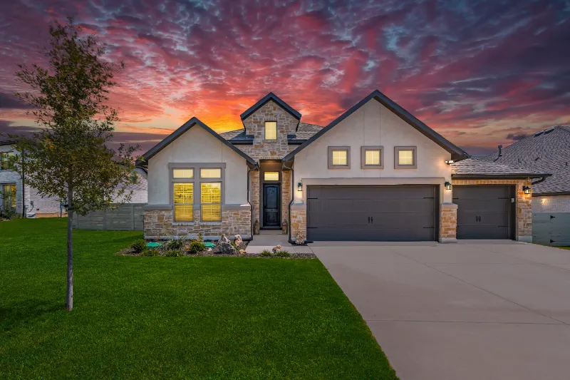 A modern, single-story house with stone accents and a three-car garage is shown at sunset. The sky is vibrant with red and purple clouds, and the front lawn is neatly landscaped with a small tree and flower beds.